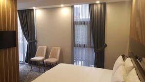 A hotel room with a bed, two chairs, a TV, and large windows with curtains. - Pozcu Otel (Mersin)