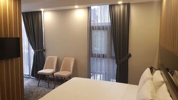 Una habitación de hotel con cama, dos sillas, televisor y ventanas grandes con cortinas.