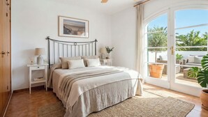 Une chambre à coucher comprenant un lit, des tables de chevet, un ventilateur de plafond, une fenêtre donnant sur un espace verdoyant et une porte.