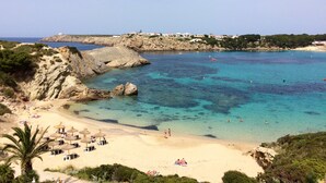 Une plage côtière aux eaux turquoise et limpides, parsemée de rochers et bordée d’un rivage sablonneux. On y trouve des parasols et des chaises longues, et quelques personnes profitent de la plage.