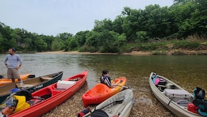 A row of kayaks and a paddleboard on a pebbly shore by a river, with a person standing nearby. - Large Bell Tent on the Kings River! (Eureka Springs)