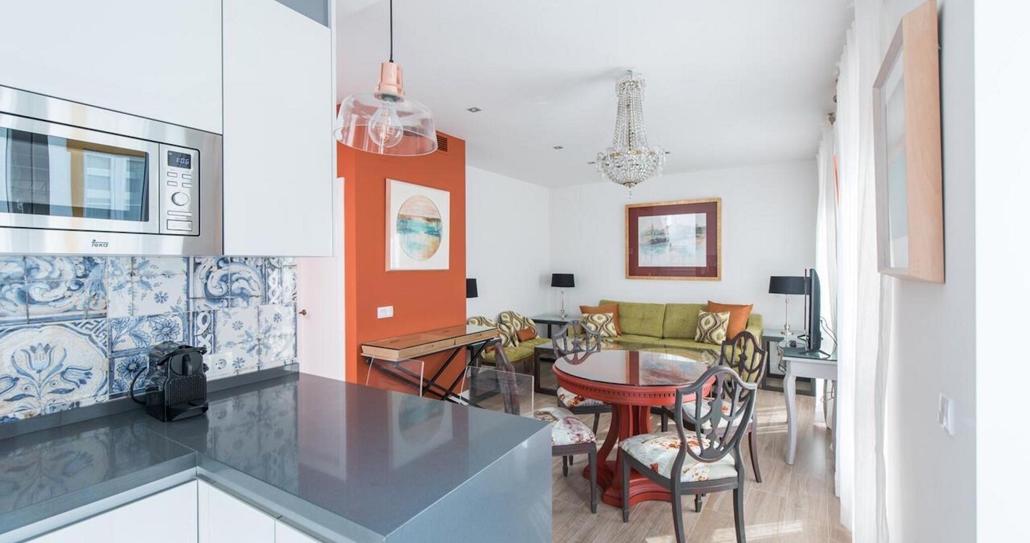 Une cuisine moderne équipée d’un four à micro-ondes Takka, un coin repas avec une table ronde et des chaises, et un salon avec un canapé et un lustre.