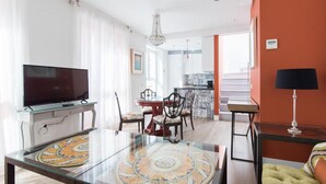 A modern living room with a flat-screen TV, a glass coffee table with a decorative center, a dining area with a round table and chairs, a kitchen with a tiled backsplash, and a staircase leading to an upper level. - Tourist apartment, located in the center of Seville (SEVILLA)