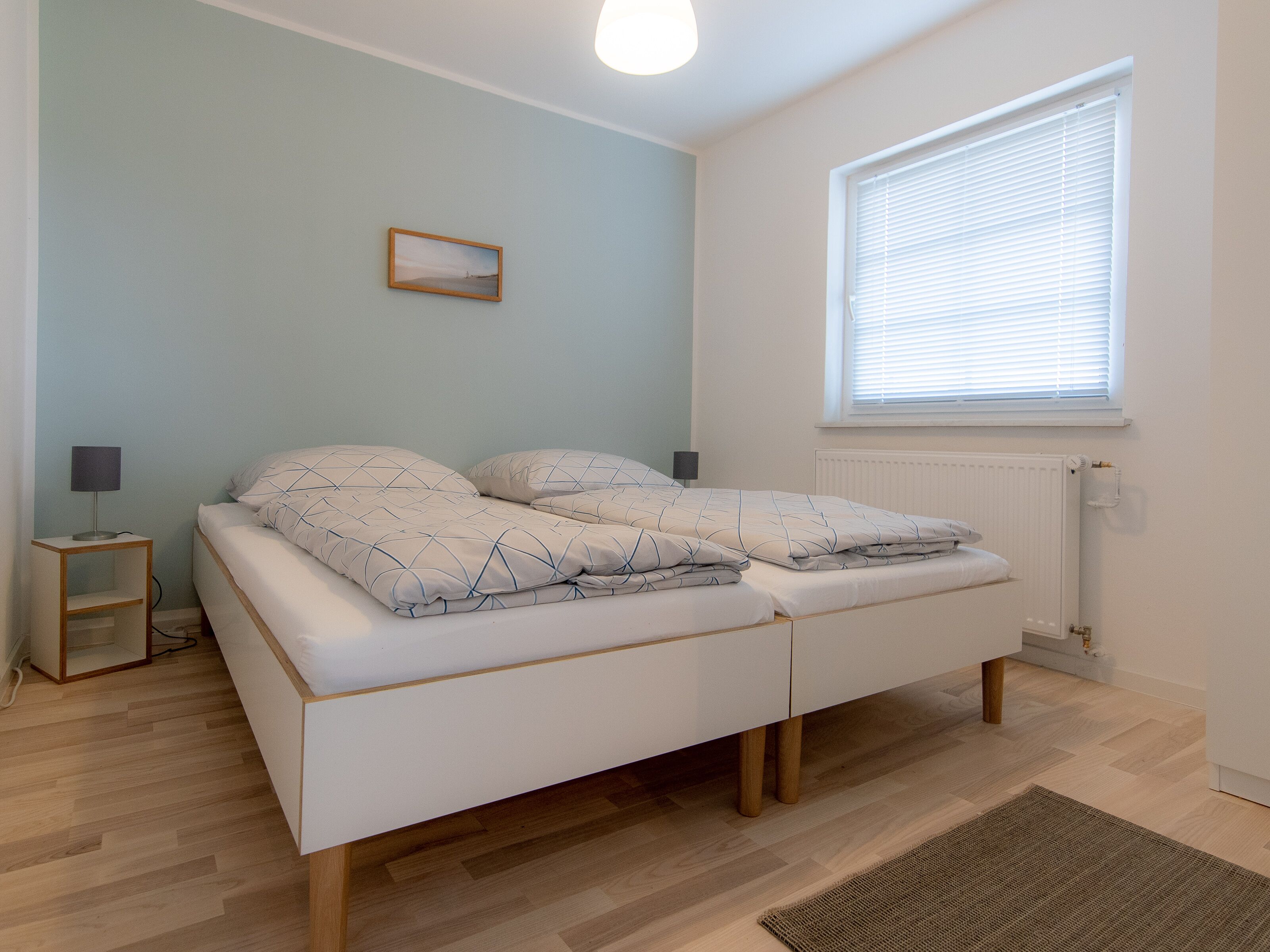 Une chambre à coucher avec un lit, des tables de chevet, une fenêtre avec des stores, un radiateur et un tableau accroché au mur.