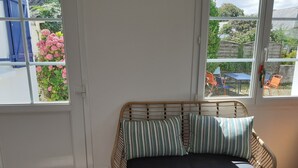 A corner room with a wicker sofa and striped pillows, a white door, and large windows overlooking a garden with chairs and tables.