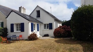 A white house with blue shutters, a chimney, and a small garden with a table and chairs.