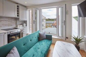 A modern kitchen with white cabinets, a teal sofa, and a view of the street through open doors.