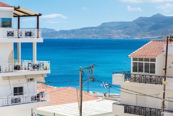 A coastal building with balconies overlooking the sea, a clear sky, and distant mountains.