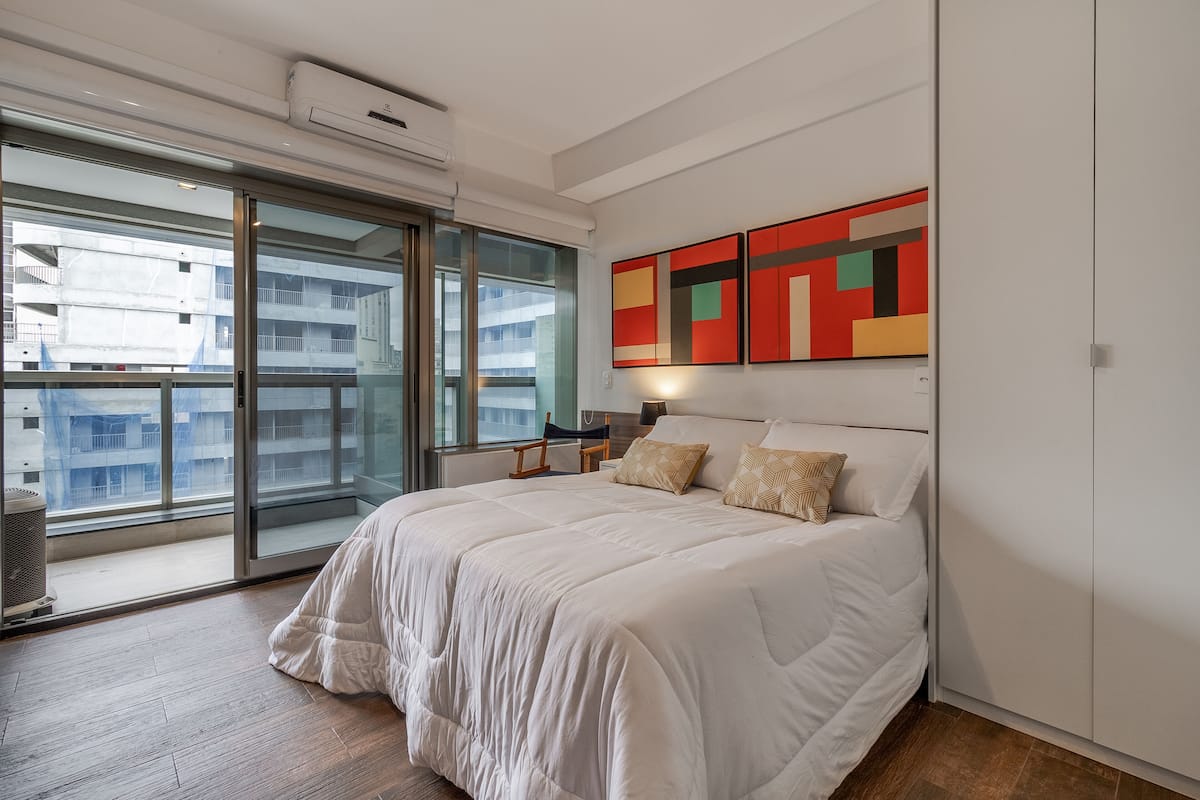 A modern bedroom with a large bed, a sliding glass door to a balcony, and a colorful abstract painting on the wall.