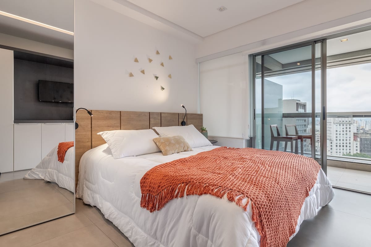 A modern bedroom with a large bed, a TV mounted on the wall, and a balcony with a city view.