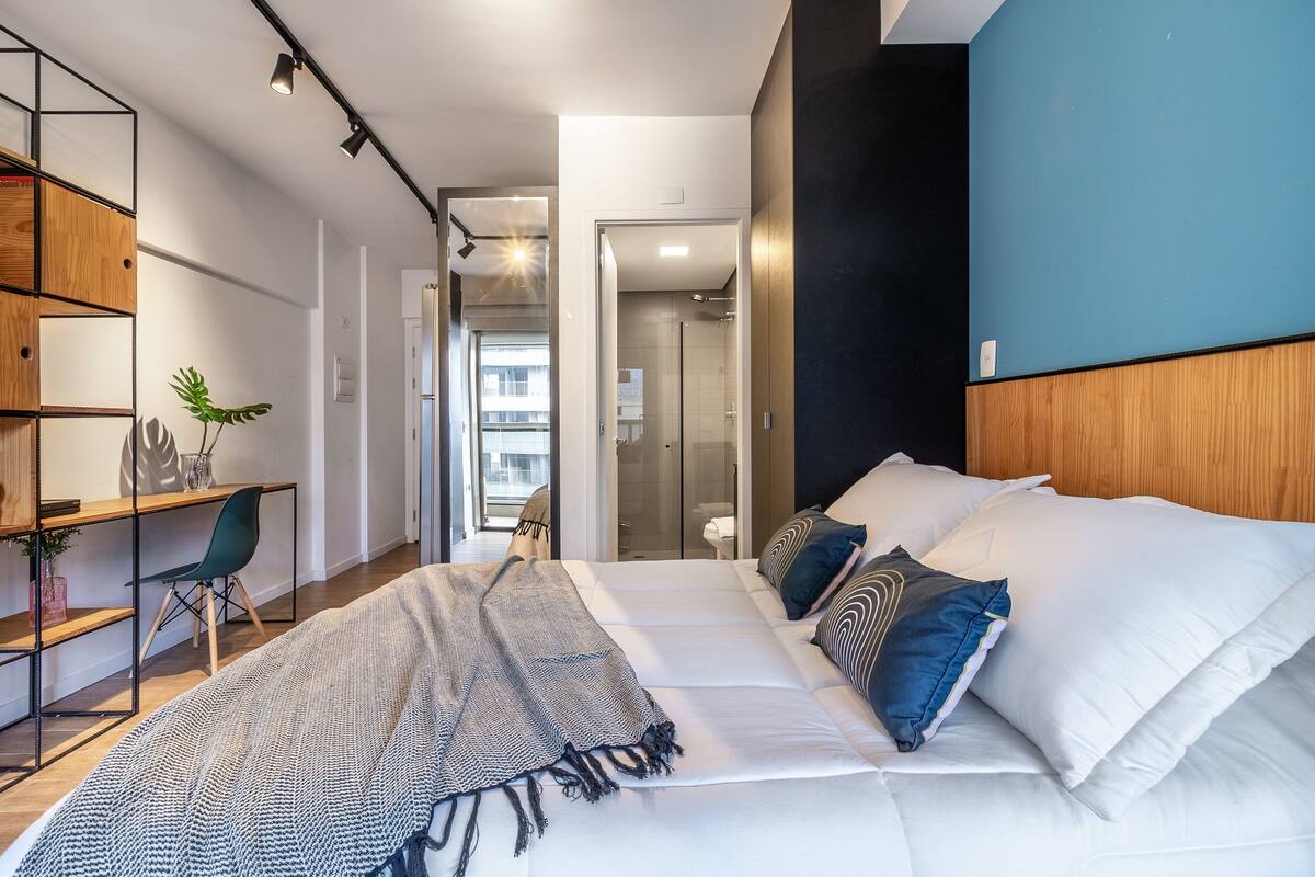 A modern bedroom with a large bed, a desk, and a chair. There is a bookshelf and a plant on the desk.