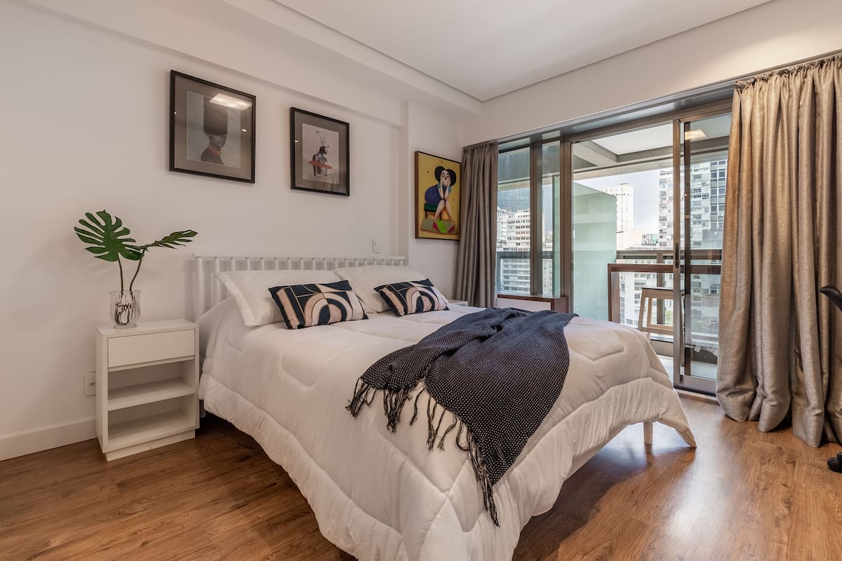 A modern bedroom with a large bed, white nightstands, and a view of the city through floor-to-ceiling windows.