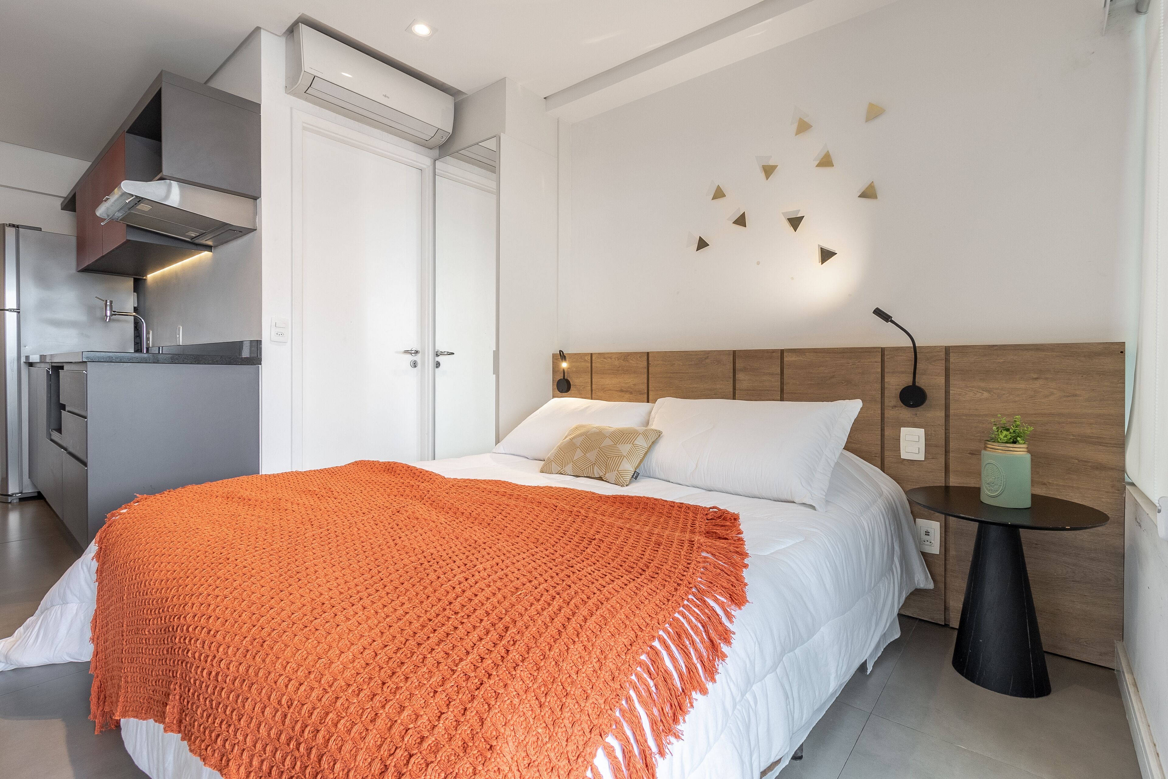 Une chambre moderne avec un lit, du linge de lit blanc, un plaid orange, une table de chevet avec un vase vert et un luminaire fixé au mur.