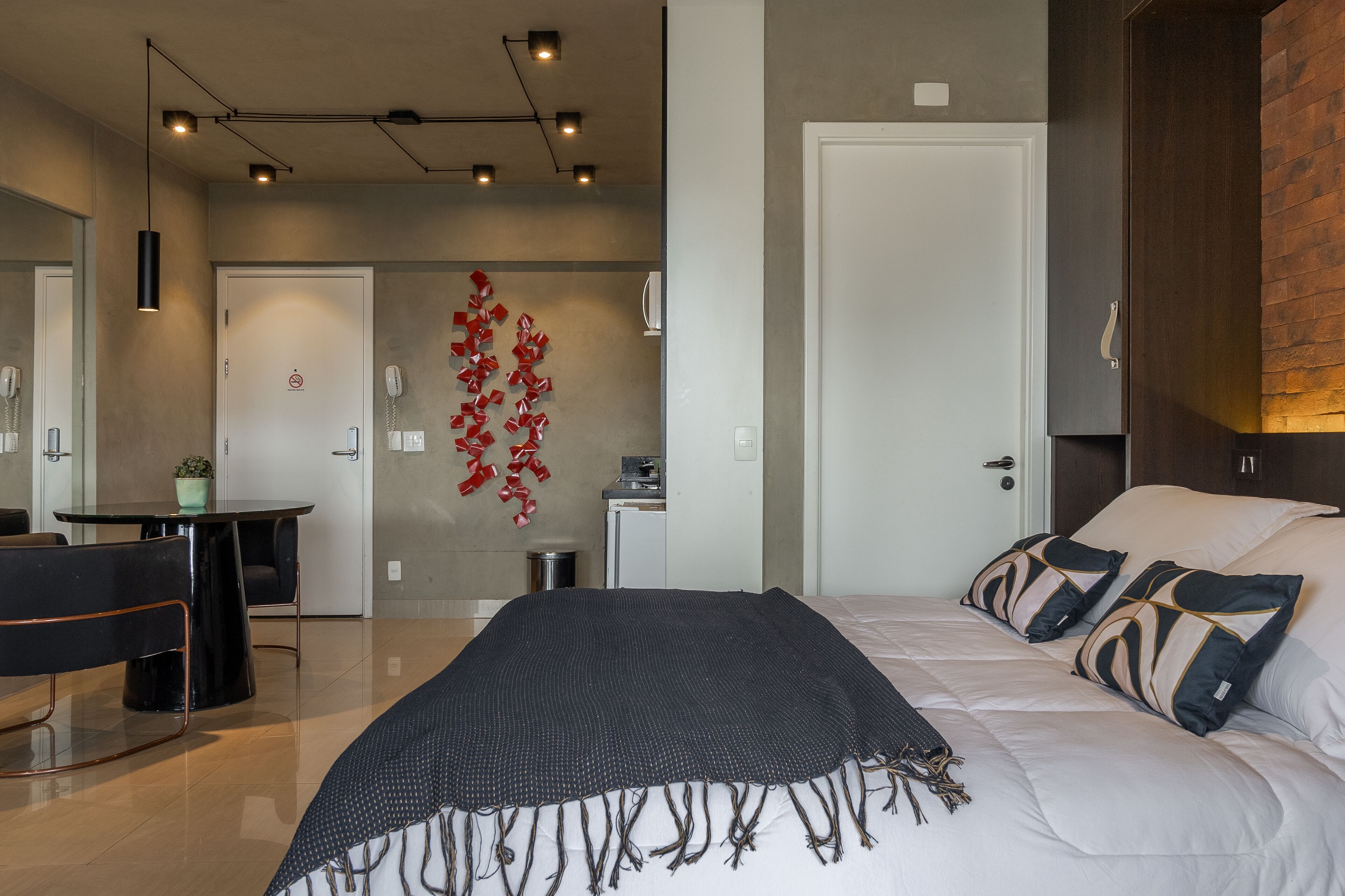 Una habitación de hotel moderna con una cama grande, un escritorio y una silla. Se observa una puerta con un adorno rojo en la pared y una pequeña mesa con una planta.