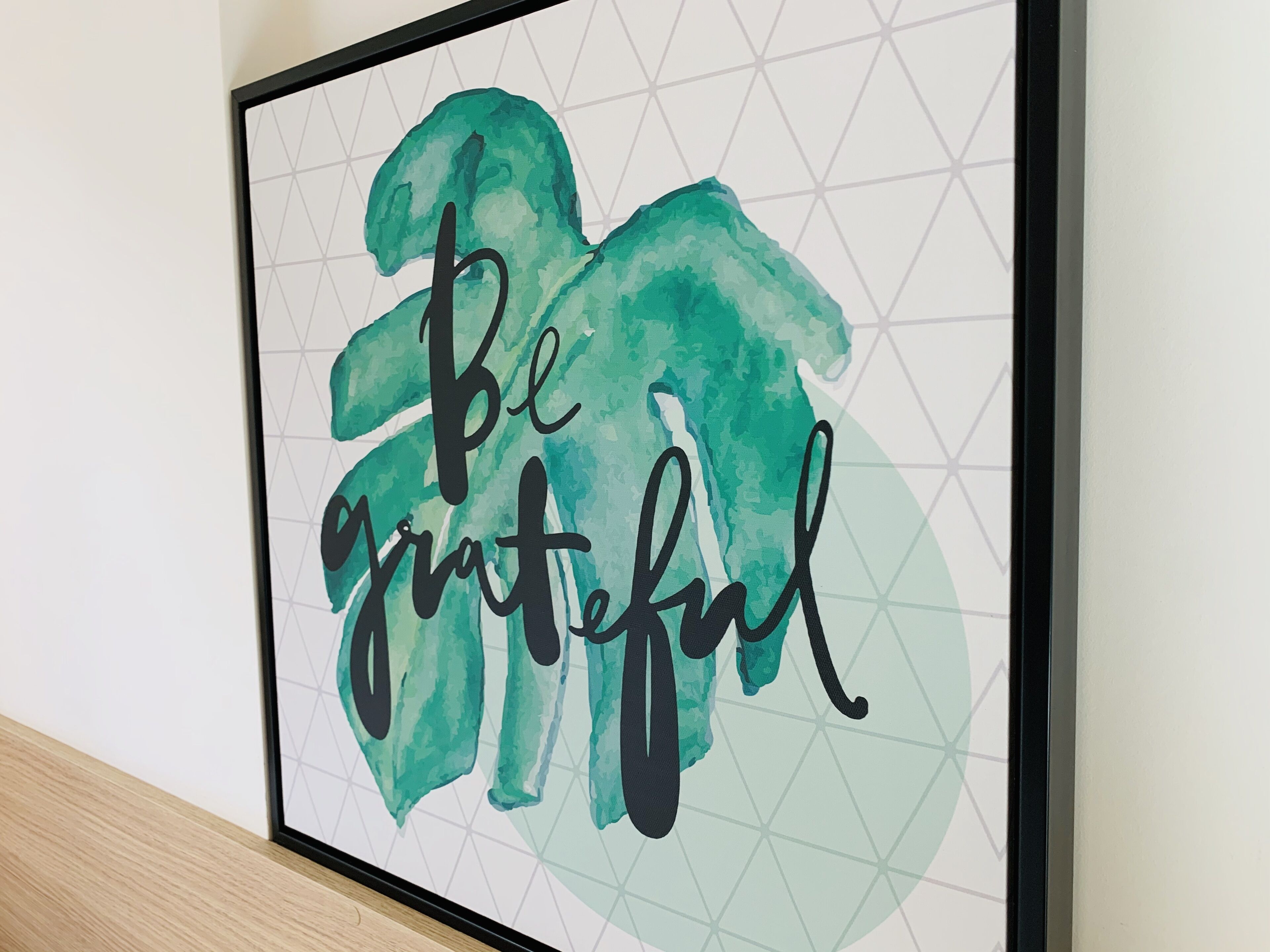 A framed artwork with the phrase "Be grateful" in a stylized font, set against a geometric background.