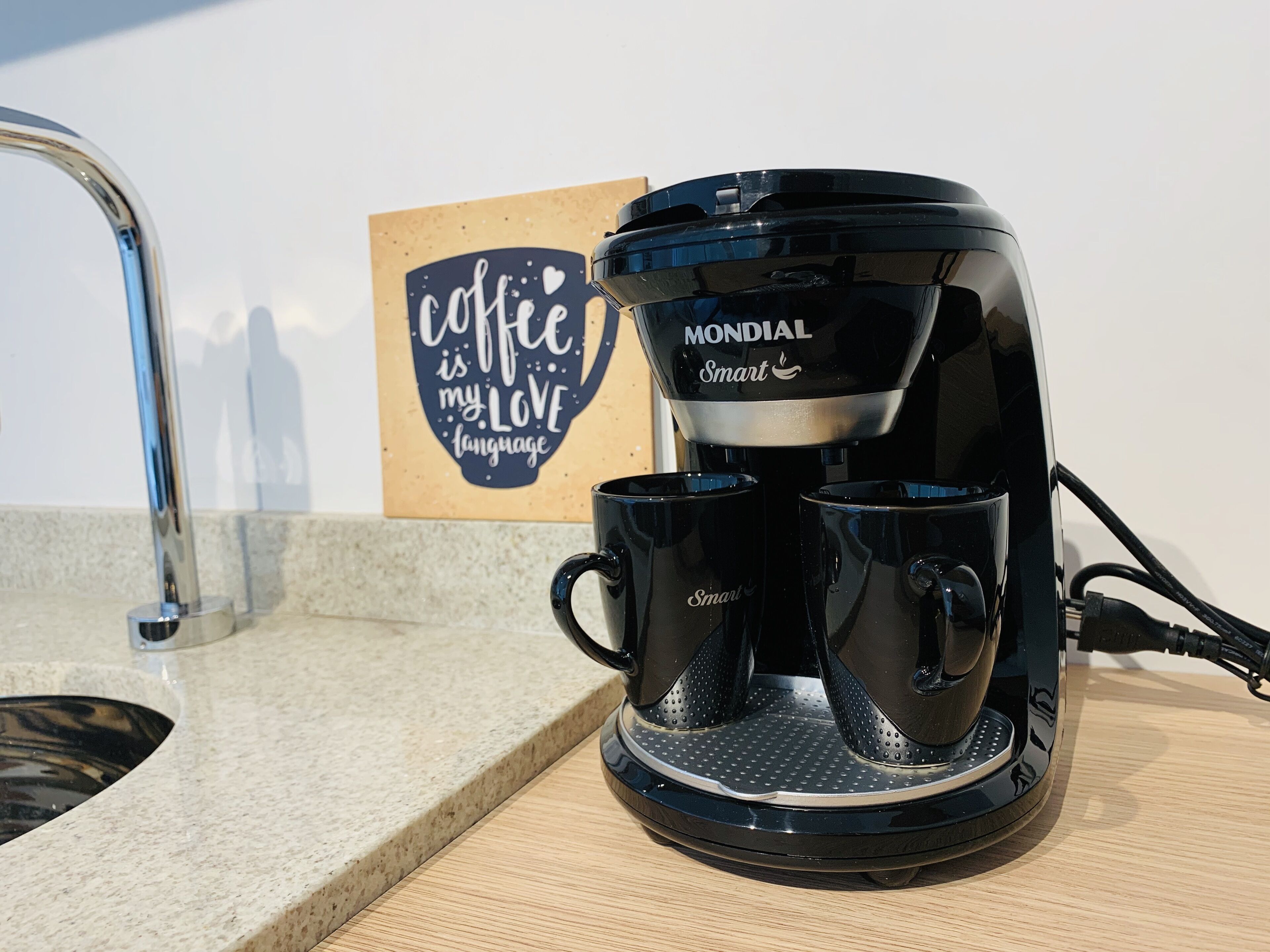 A coffee maker with two cups on a countertop, a sink, and a wall with a coffee-themed sign.
