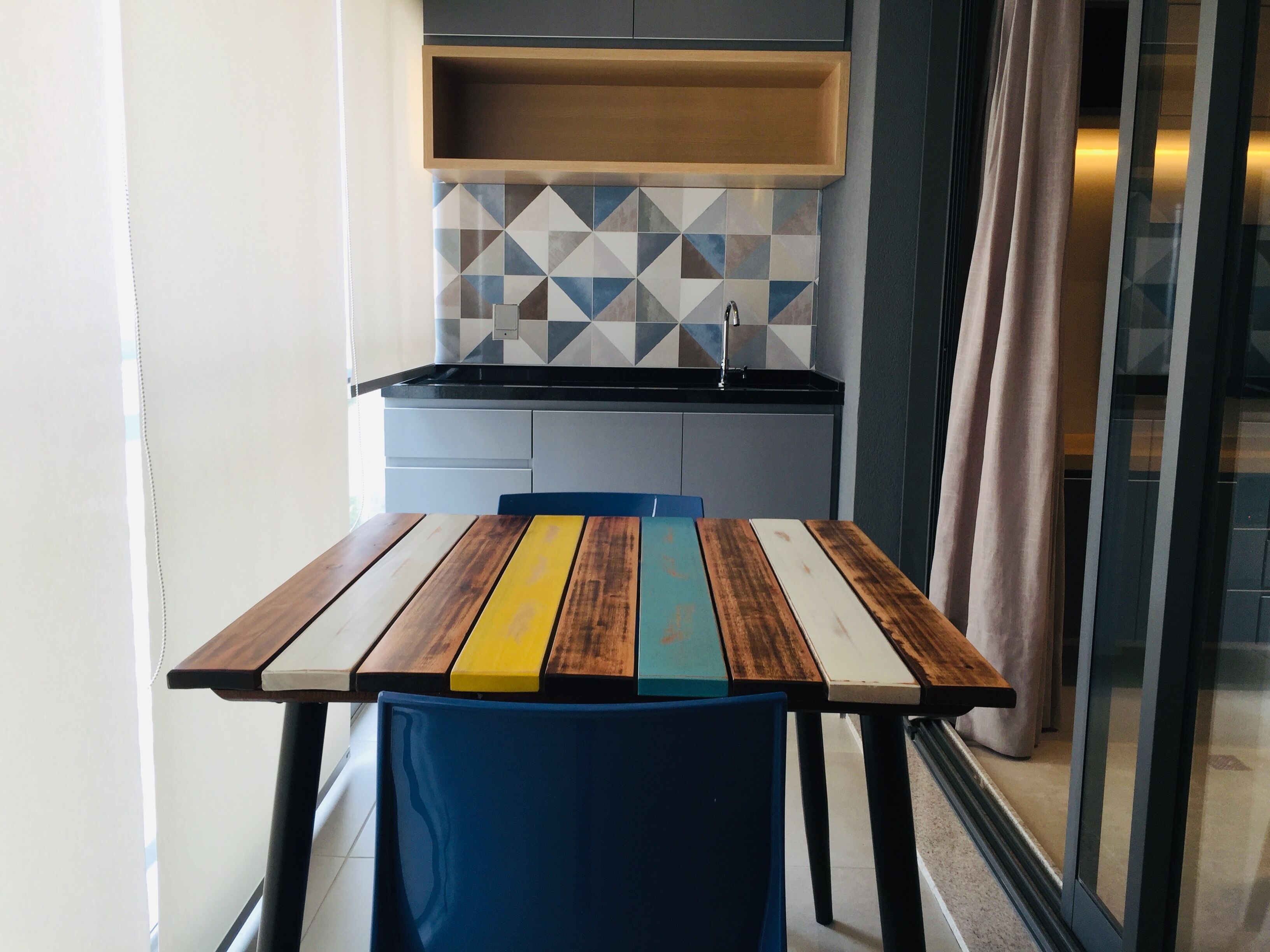 A compact kitchen with a modern design, featuring a geometric tile backsplash, a wooden table with colorful stripes, and a blue chair.