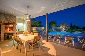 Un área de comedor al aire libre con mesa y sillas, una chimenea y una piscina al fondo.