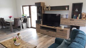 Un salon moderne avec une table basse en bois, un canapé bleu, un téléviseur posé sur un meuble en bois, et un coin repas comprenant une table et des chaises.