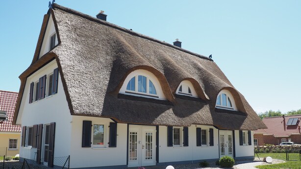 Ett traditionellt hus med ett brant, halmtäckt tak, många fönster och en välskött trädgård med stenar och blommor.