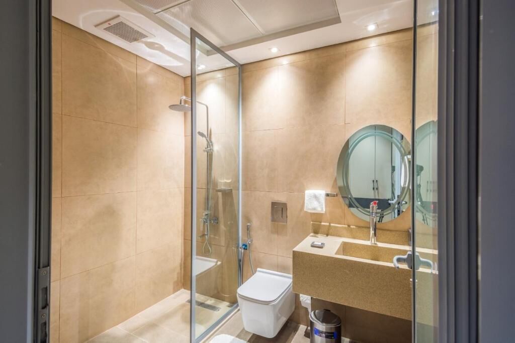 A modern bathroom with a glass shower enclosure, a white toilet, and a beige sink with a round mirror.