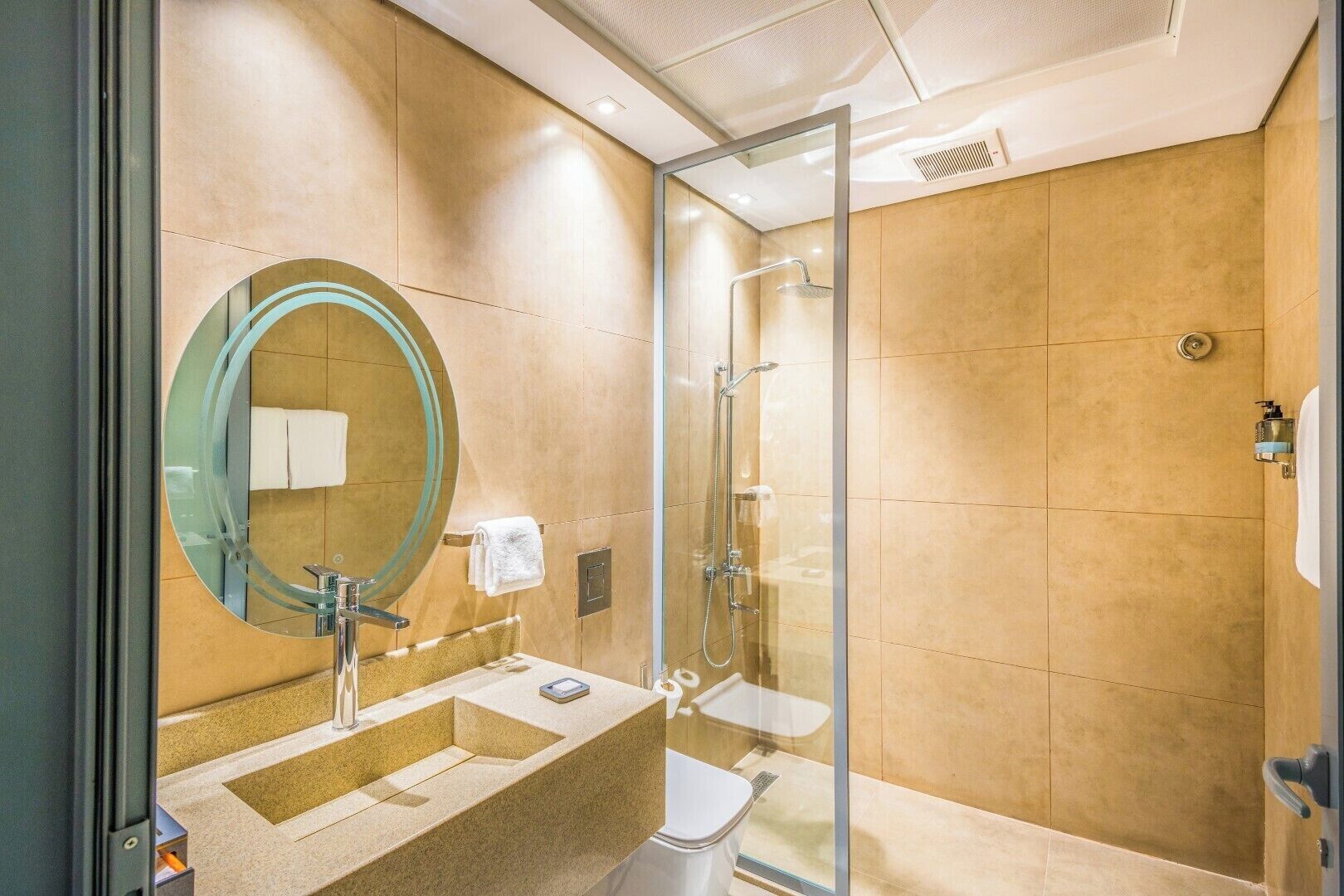 A modern bathroom with a glass-enclosed shower, a round mirror, and a beige-tiled vanity.