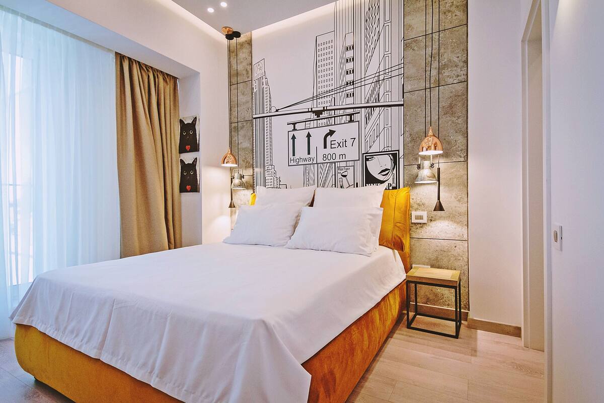 A modern hotel room with a large bed, a cityscape mural on the wall, and a small table with a lamp.