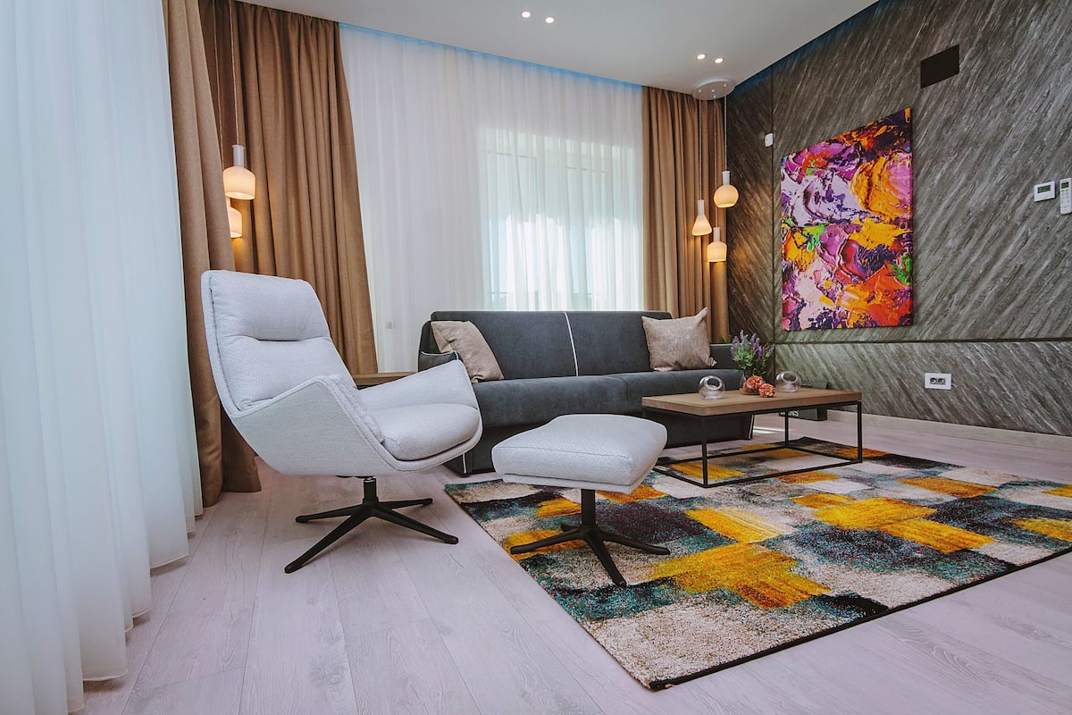 A modern living room with a sofa, armchair, and ottoman, a colorful abstract painting on the wall, and a patterned rug.