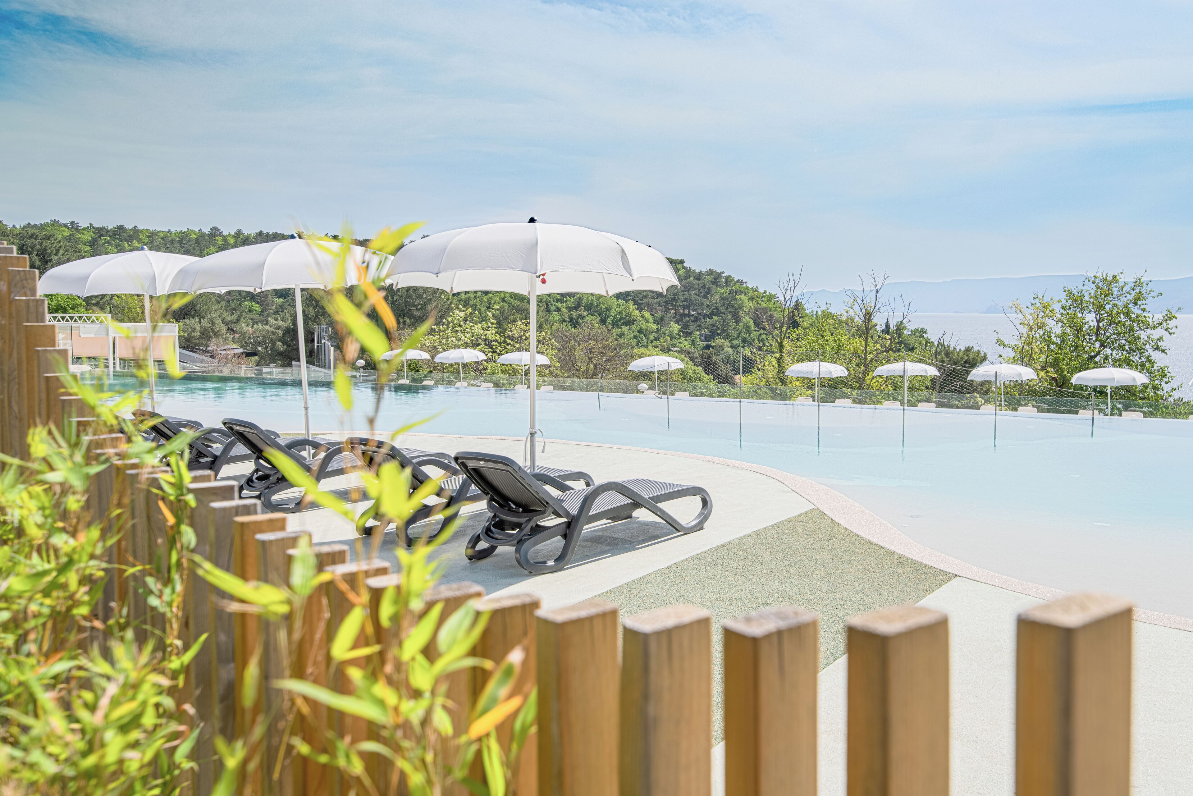 Un espace aménagé au bord de la piscine, avec des chaises longues et des parasols, entouré d’une clôture en bois et de verdure.