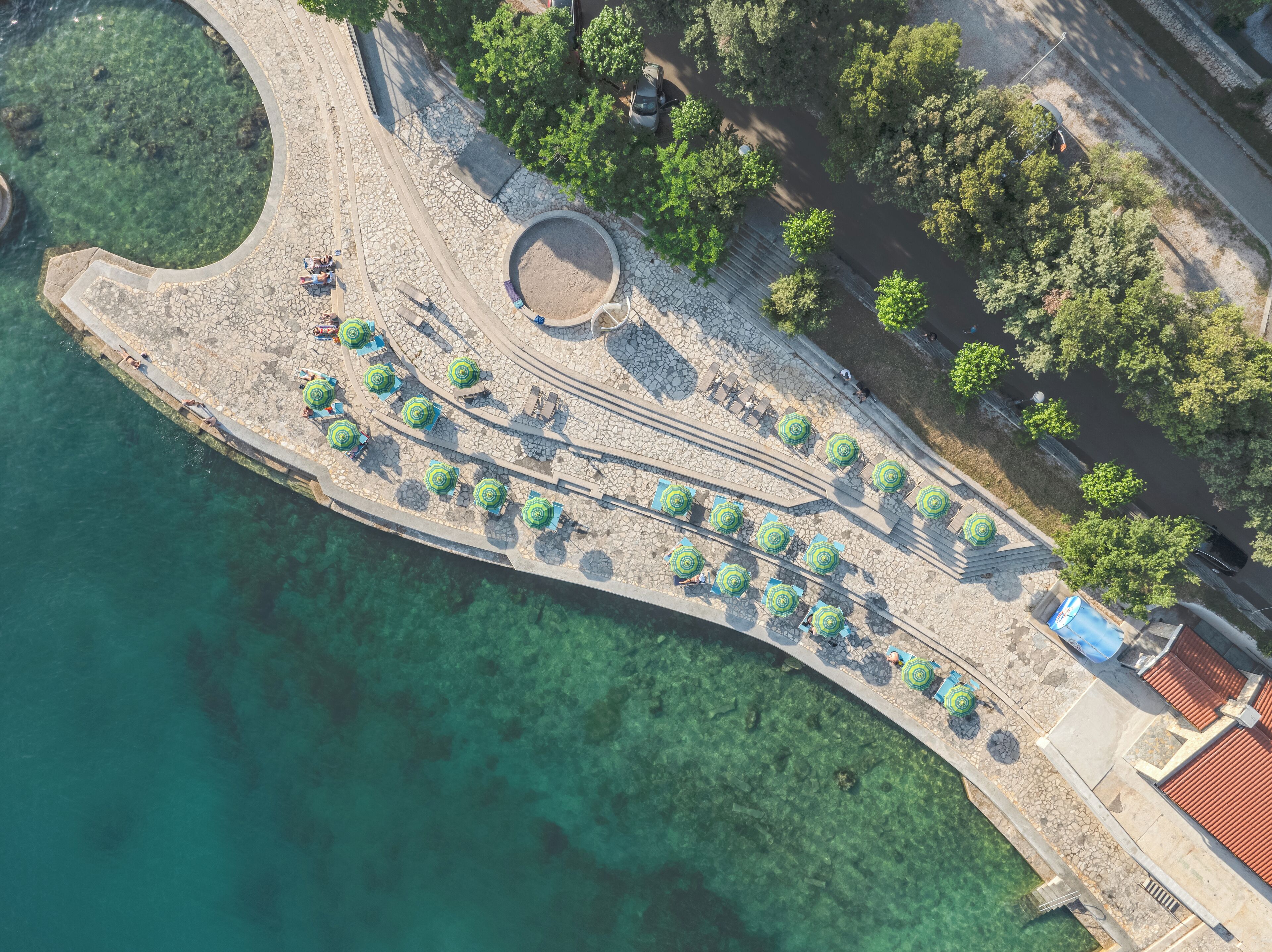 An aerial view of a coastal promenade with clear waters, stone pathways, and green umbrellas.
