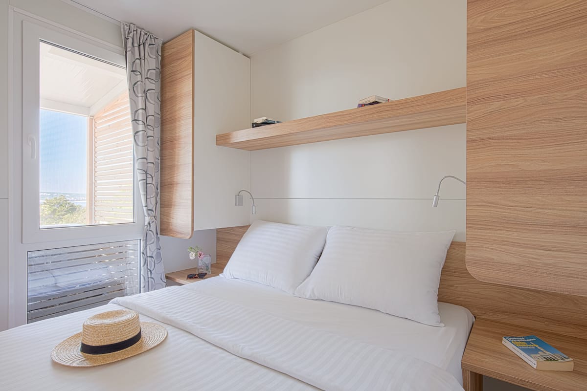 Habitación de hotel con cama, ventana con persianas, un sombrero sobre la cama y una estantería con libros.