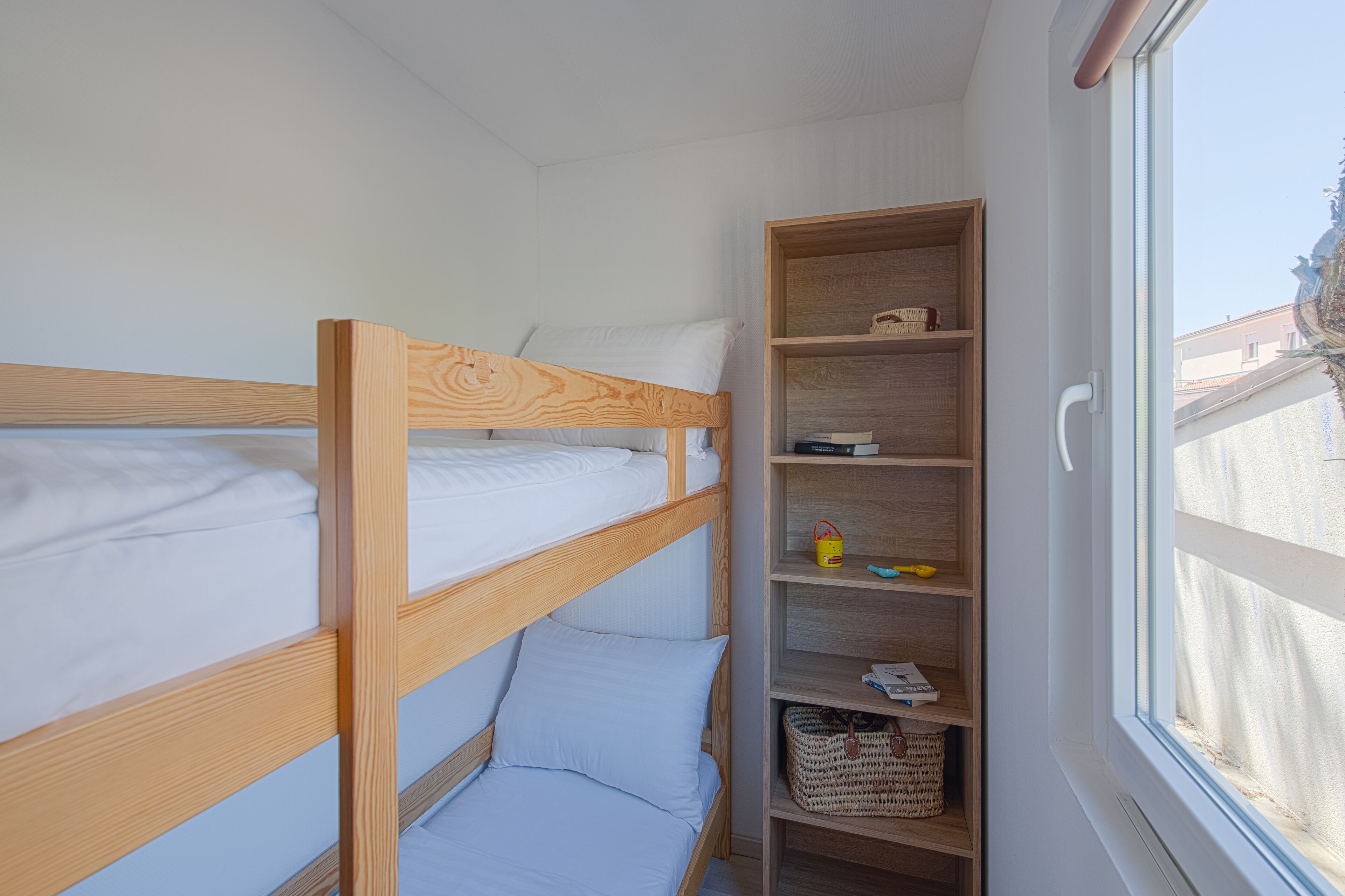 Una camera con un letto a castello, una testiera in legno, uno scaffale con libri e un cesto, e una finestra con vista sugli edifici.