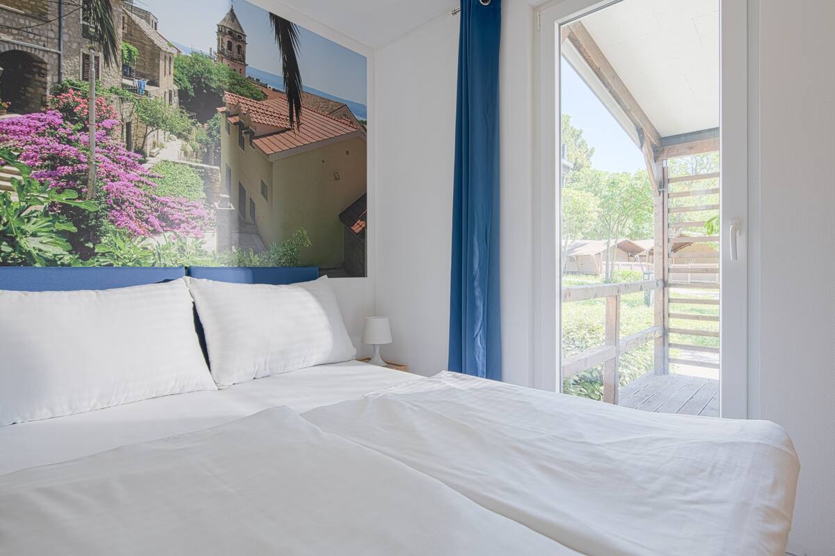 Un dormitorio con cama, almohadas blancas, una mesita de noche, una lámpara y vistas a un balcón con barandilla de madera.