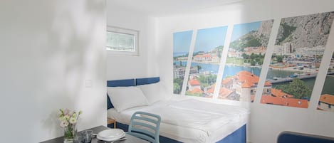 A modern room with a bed, a dining table with plates and glasses, and a view of a coastal town through a large window.