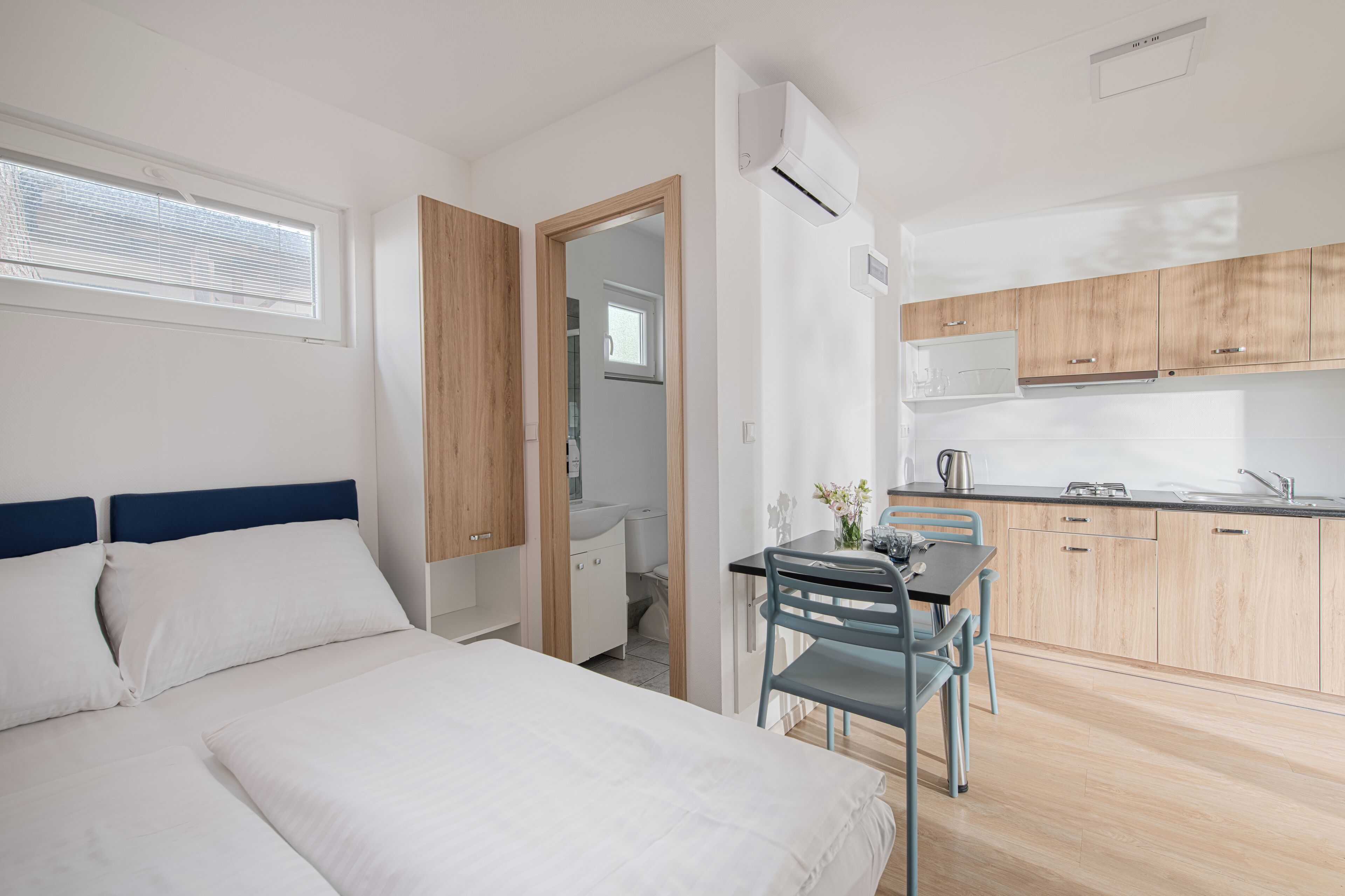 A modern hotel room with a bed, a small dining table with chairs, a kitchenette, and a bathroom visible through an open door.
