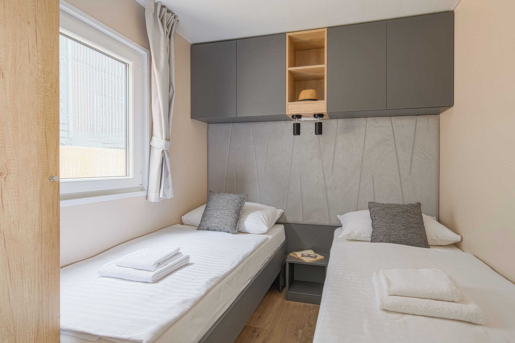 A modern hotel room with two single beds, a small bedside table, a built-in shelf, and a window with curtains.