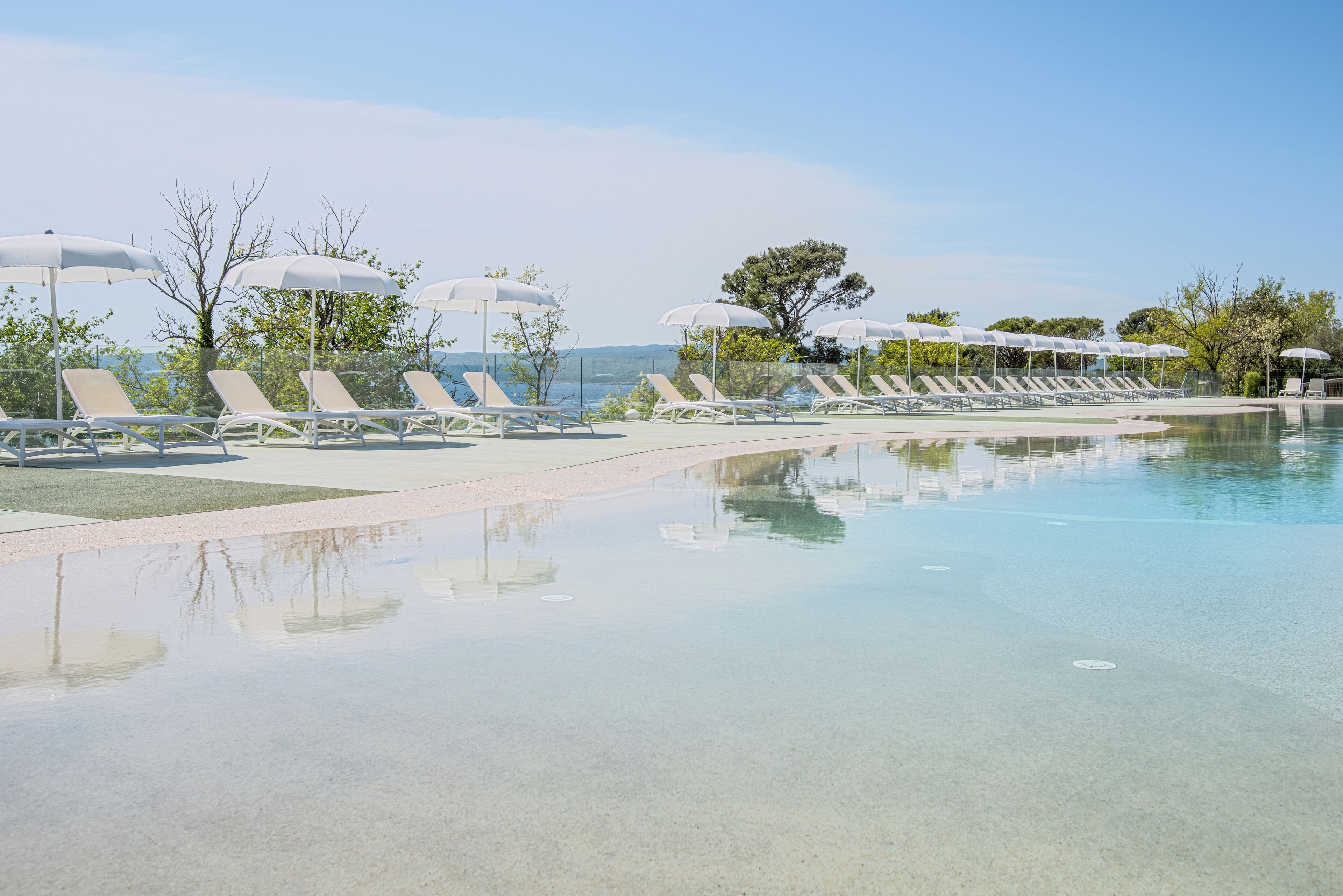 Zona de piscina con tumbonas y sombrillas, agua cristalina y vistas al paisaje.