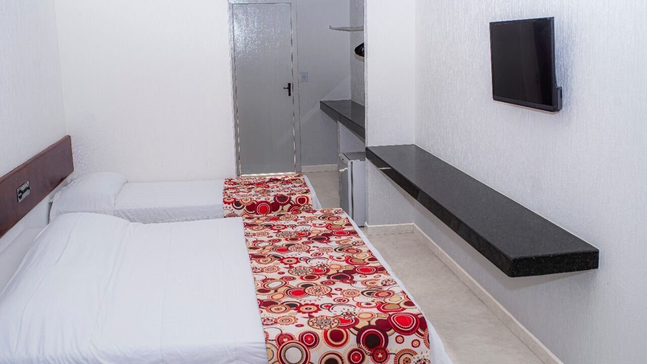 A small hotel room with two beds, a TV mounted on the wall, and a kitchenette area.