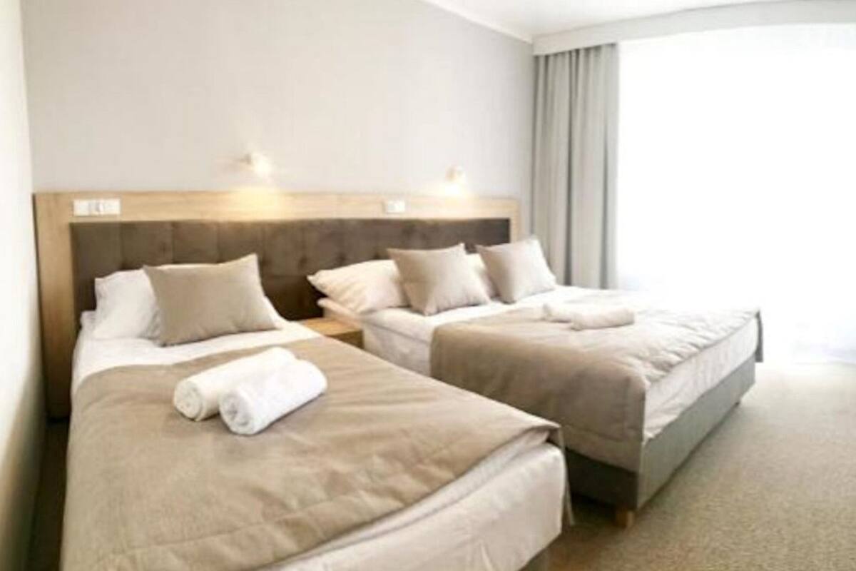 Une chambre d’hôtel avec deux lits simples, une tête de lit en bois et une grande fenêtre avec des rideaux.