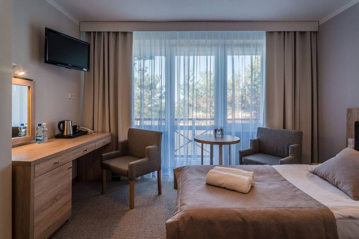 Une chambre d’hôtel comprenant un lit, deux fauteuils, un bureau avec une télévision, une petite table et une grande fenêtre avec des rideaux.