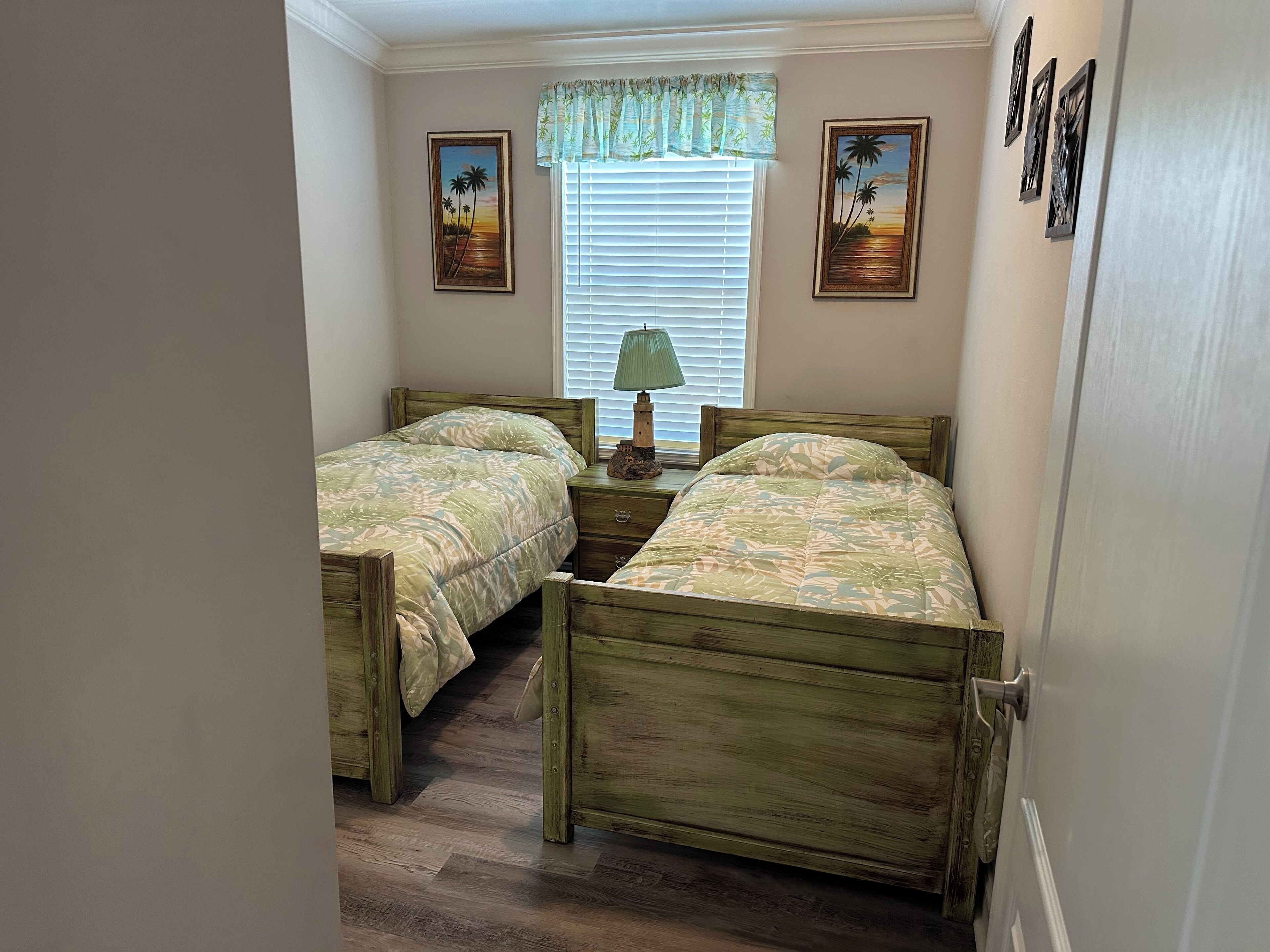 A bedroom with two wooden beds, a window with blinds, a lamp, and two framed pictures on the wall.