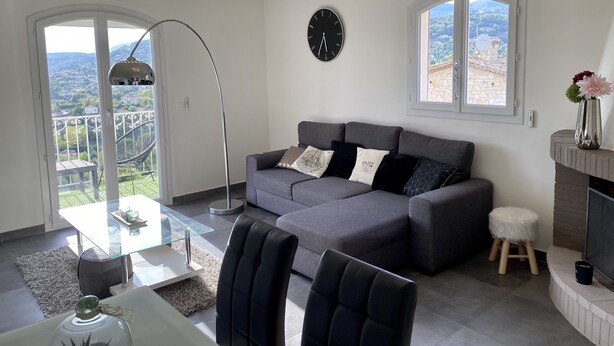 Una sala de estar moderna con un sofá gris, cojines blancos y negros, una mesa de centro de vidrio y una silla negra. Hay un reloj redondo en la pared y una ventana con vistas al campo.