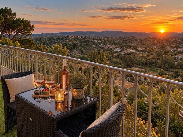 Un balcón con mobiliario de mimbre, una mesa dispuesta con copas de vino, una botella y aperitivos, con vistas a un paisaje escénico al atardecer.
