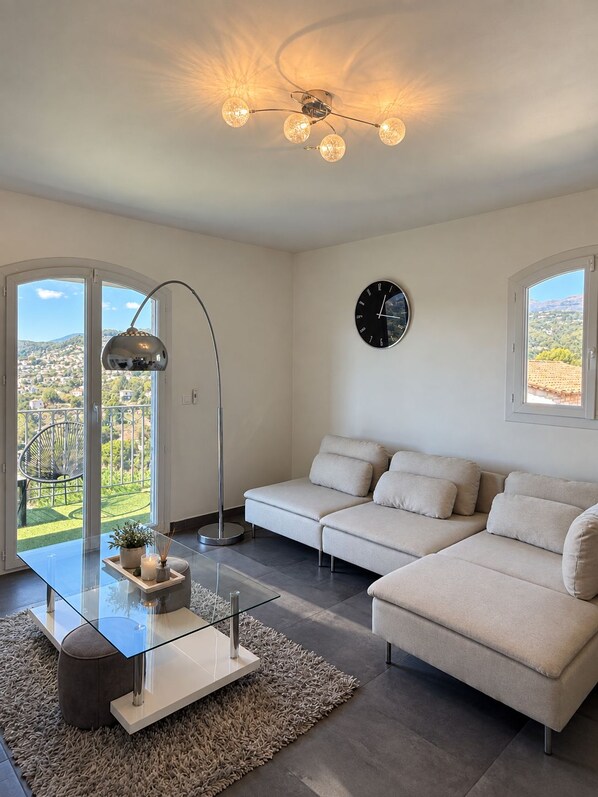 Ein modernes Wohnzimmer mit einem Glastisch, beigefarbenen Sofas, einer Stehlampe und Blick auf eine grüne Landschaft durch große Fenster.