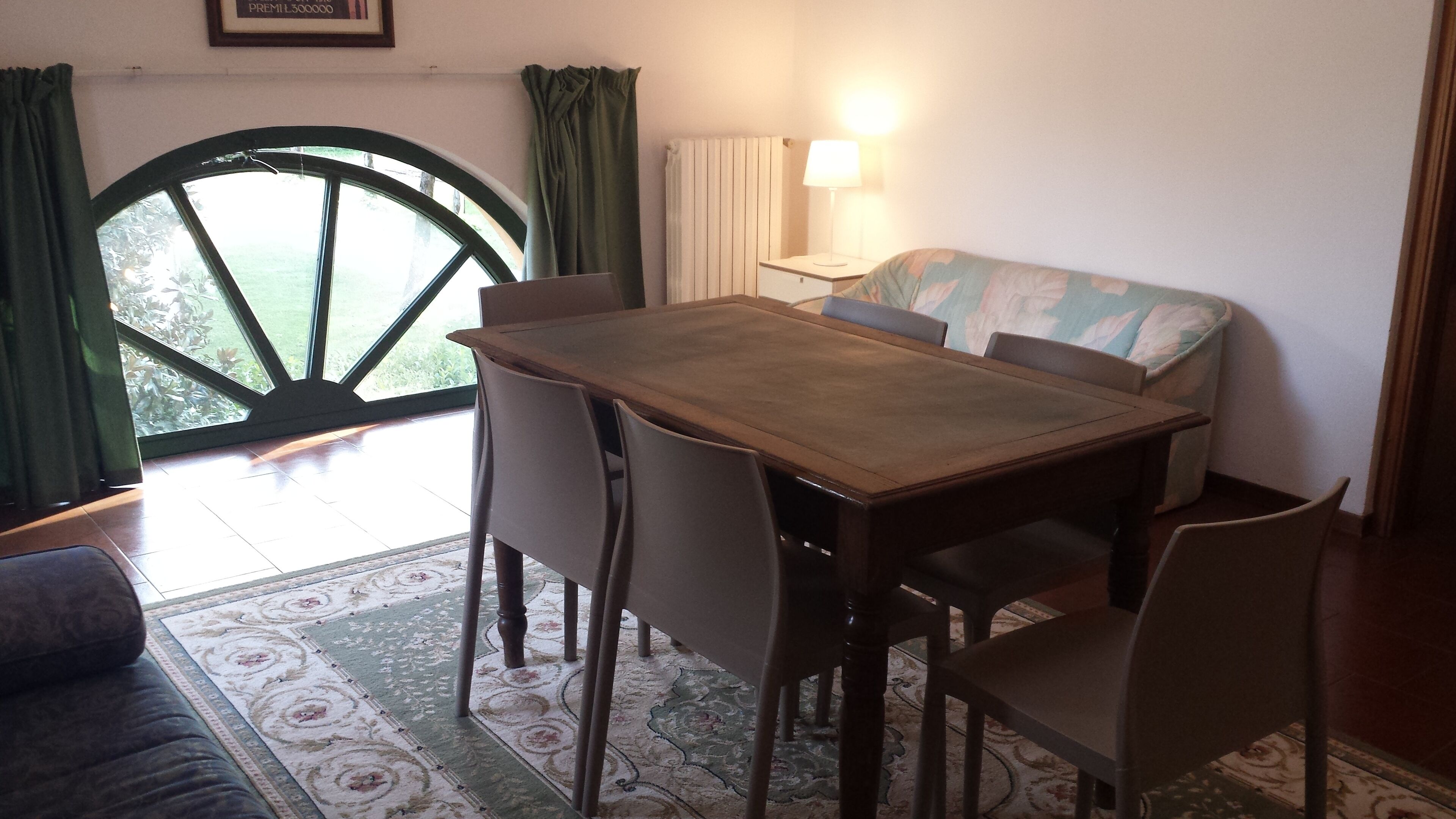 Un área de comedor con una mesa y sillas de madera, un sofá, una lámpara y vistas al exterior a través de un ventanal con cortinas.