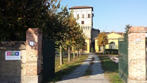 A pathway leading to a building with a tower, surrounded by trees and a brick wall with a gate. - Palazzo Barbò (Torre Pallavicina)