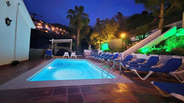 A swimming pool with lounge chairs and a pergola, surrounded by palm trees and a hillside.