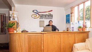 Un mostrador de recepción de madera con una computadora portátil, una planta en maceta y un cartel que dice 'Summita Hotel & Restaurant'.