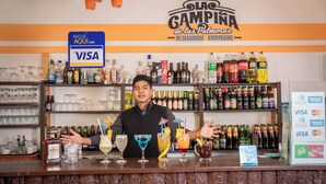 Zona de bar con un letrero que indica pago con Visa, diversas bebidas alcohólicas y una pizarra de menú.