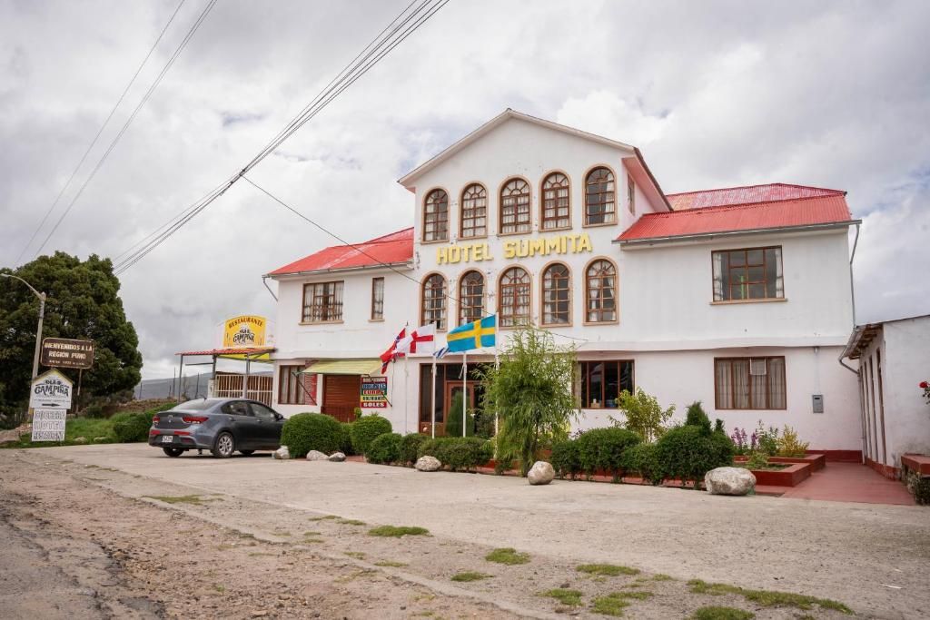 Un bâtiment à deux étages, avec un toit rouge et une enseigne indiquant « HÔTEL SUMMITA ». On peut voir des drapeaux, une voiture garée et une enseigne de restaurant au premier plan.